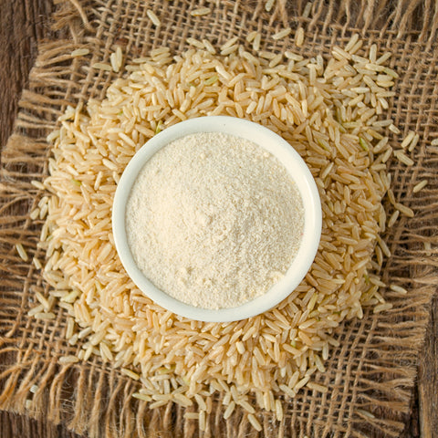 Bowl of organic brown rice flour surrounded by organic raw brown rice on a rustic background