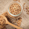 Glass jar filled with dried raw buckwheat groats on a textured surface with a wooden spoon.