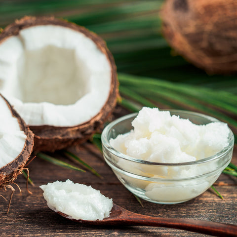 Organic coconut oil in a glass bowl with a wooden spoon and halved coconut on a wooden surface