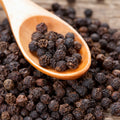 Wooden spoon filled with organic black peppercorns surrounded by more peppercorns on a wooden surface
